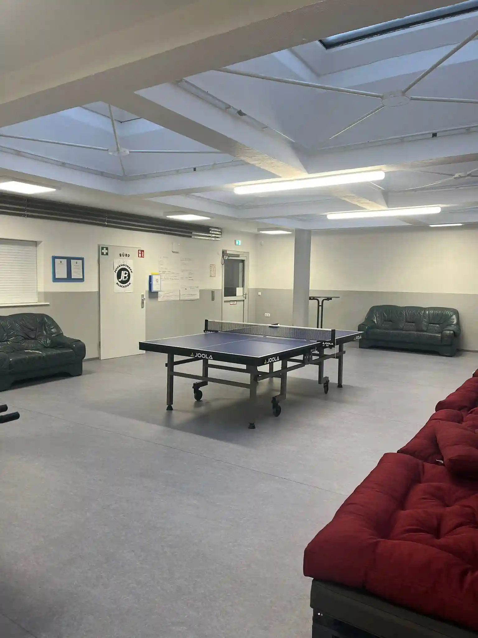 In der Mitte eines geräumigen Raums mit Sofas, Deckenbeleuchtung und Aushängen an der Wand steht eine Tischtennisplatte.