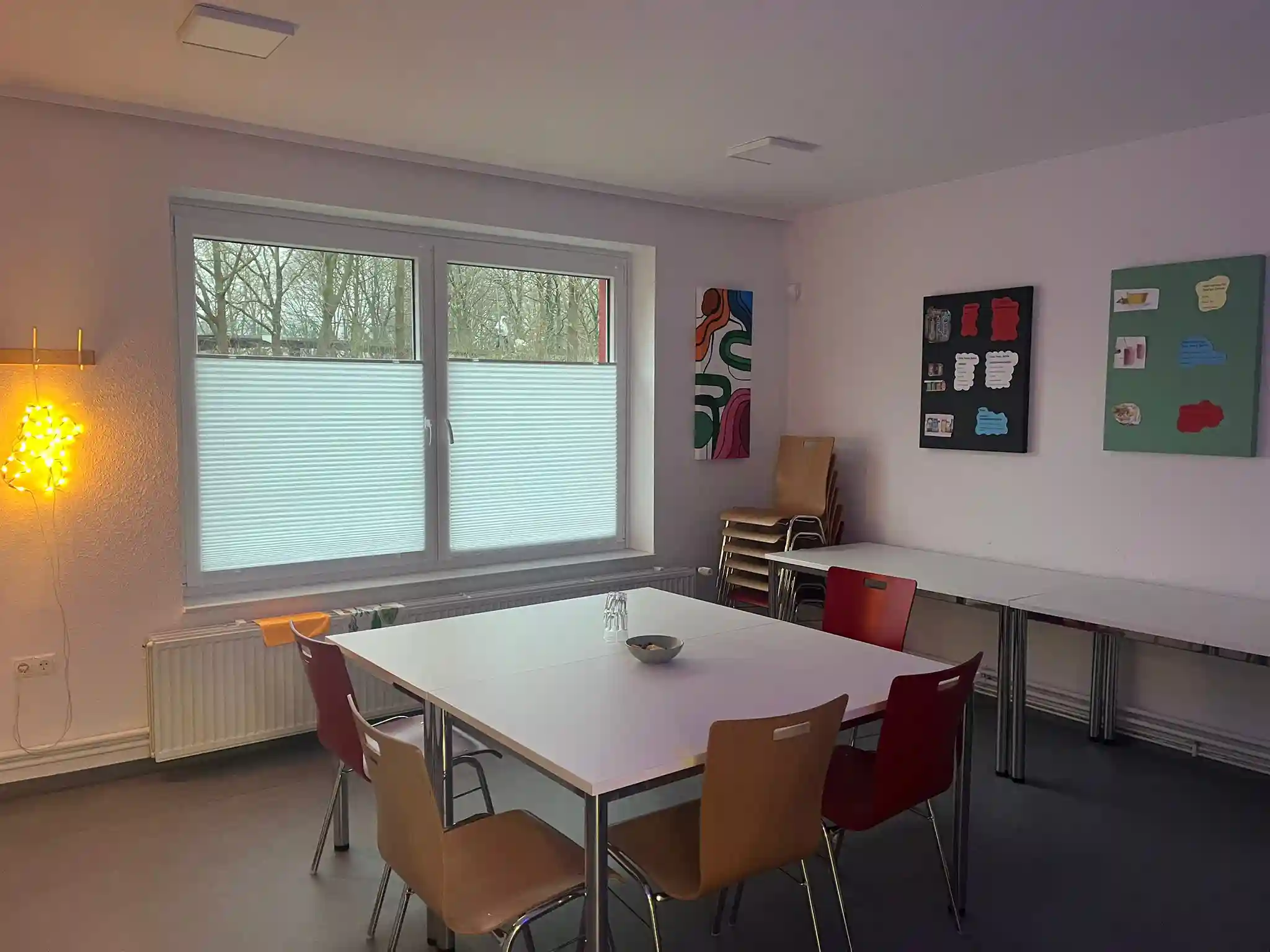 Ein kleiner Raum mit einem Tisch, verschiedenen Stühlen, in der Ecke gestapelten Klappstühlen, einem Fenster mit Jalousien, Wandbildern und einer dekorativen Wandleuchte.