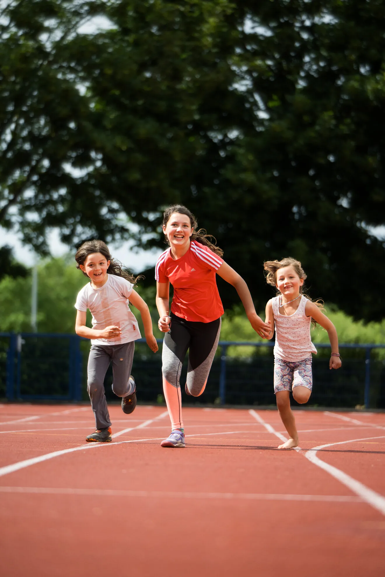 Drei Kinder laufen lächelnd gemeinsam auf einer Laufbahn im Freien, im Hintergrund sind Bäume und Grünflächen zu sehen.