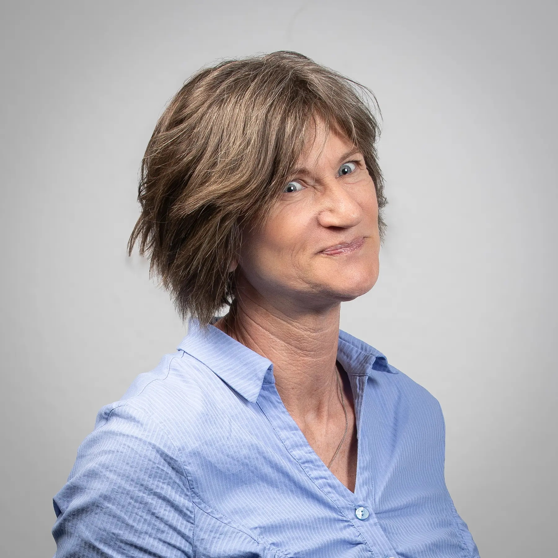 Eine Frau mit kurzen braunen Haaren, die ein hellblaues Hemd mit Knöpfen trägt, steht vor einem schlichten grauen Hintergrund und blickt mit neutralem Gesichtsausdruck leicht zur Seite.
