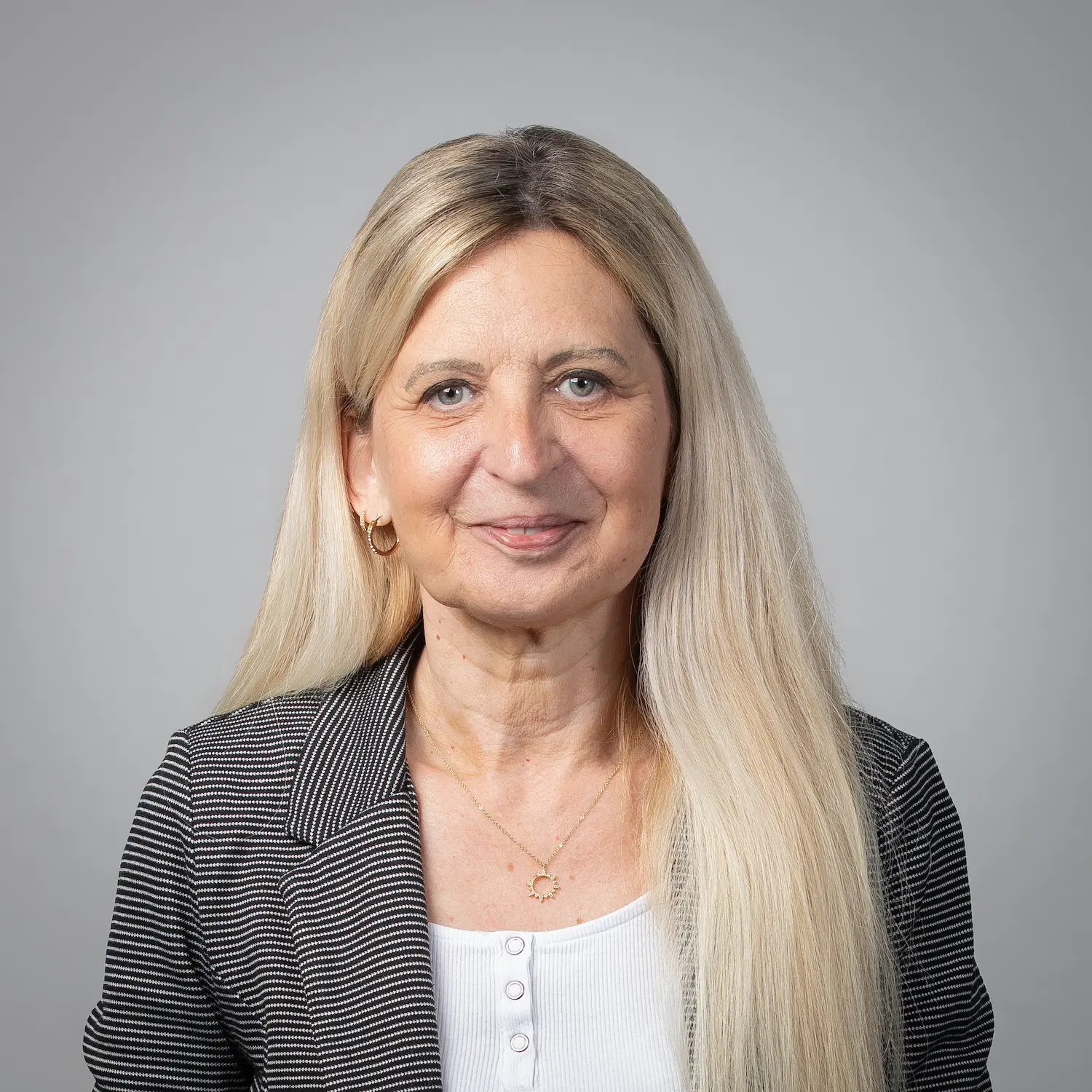 Eine Frau mit langen blonden Haaren, die einen Nadelstreifenblazer über einem weißen Hemd trägt, steht vor einem schlichten grauen Hintergrund.