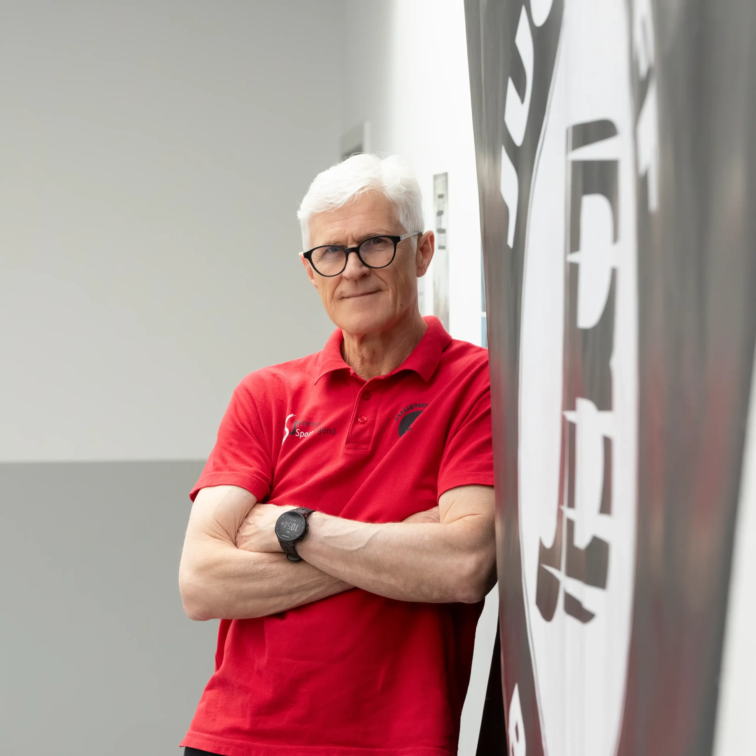 Ein älterer Mann mit weißem Haar und Brille steht mit verschränkten Armen da; er trägt ein rotes Poloshirt und eine schwarze Armbanduhr und lehnt an einer Wand, auf der ein großes abstraktes Logo zu sehen ist.
