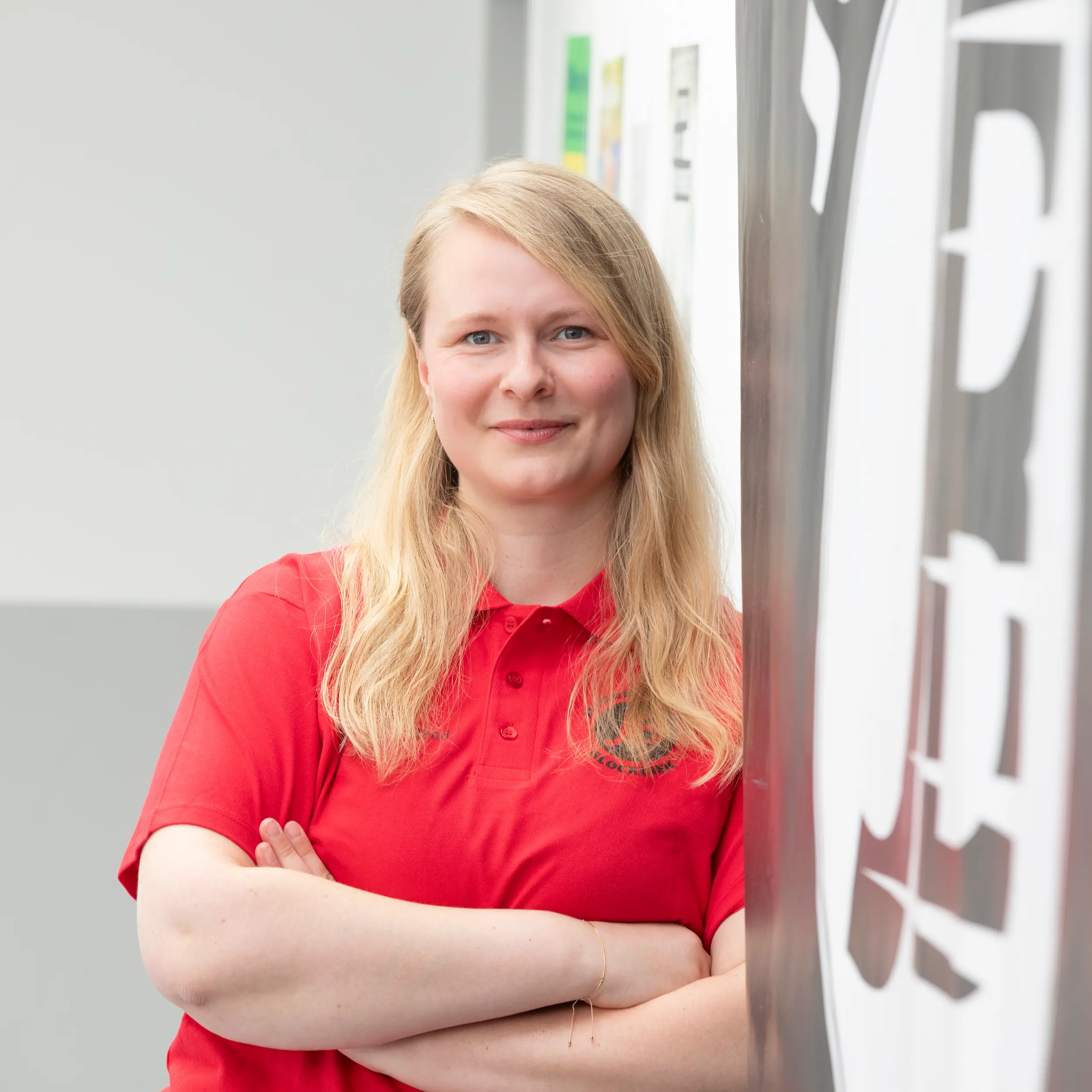Eine Person mit langen blonden Haaren, die ein rotes Poloshirt trägt, steht mit verschränkten Armen im Raum und lächelt leicht.