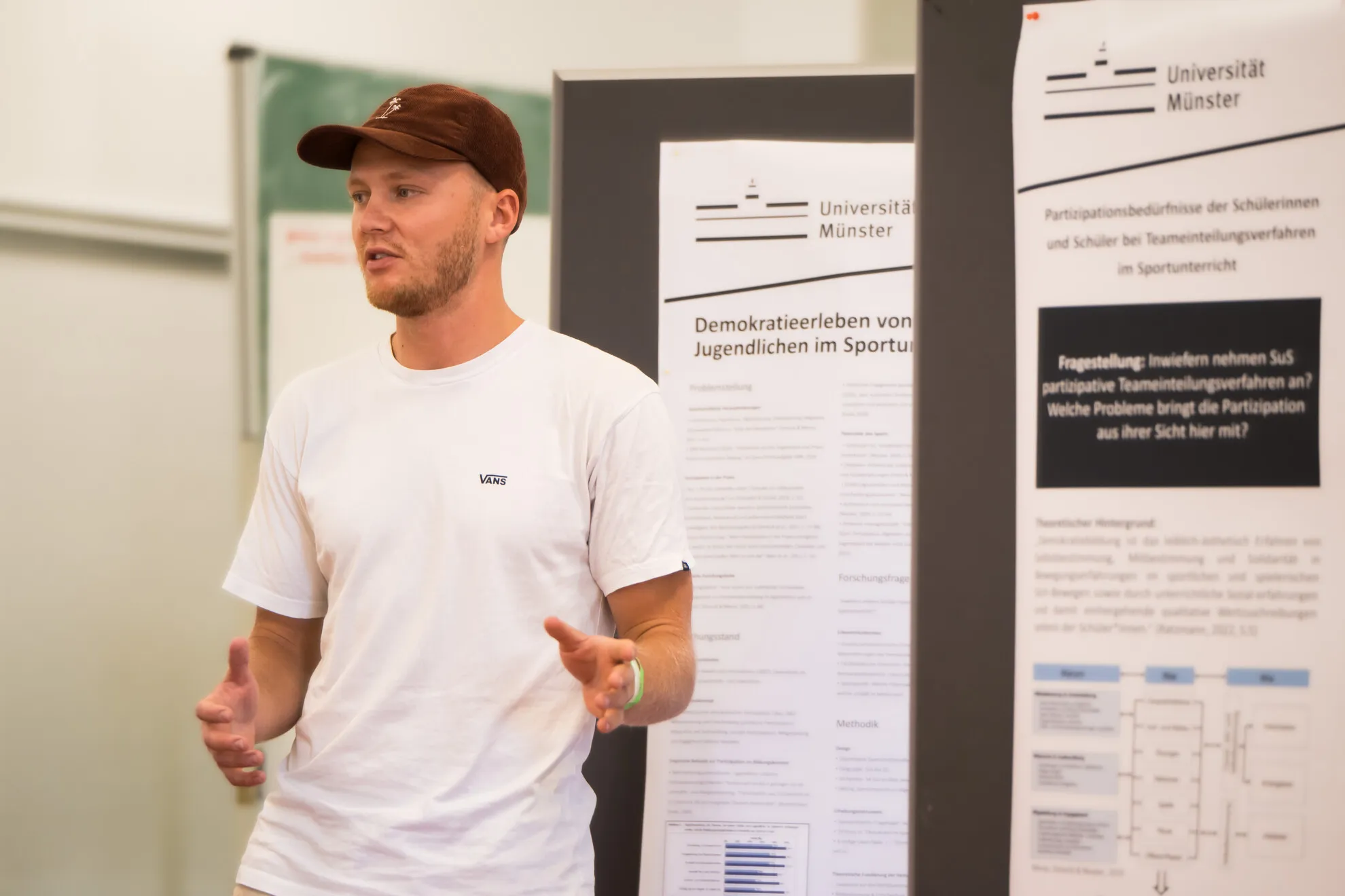 BSJ_Referent_Vortrag Ein Mann in einem weißen T-Shirt und einer Mütze spricht vor wissenschaftlichen Postern an der Universität Münster; vermutlich stellt er Forschungsergebnisse vor oder leitet eine Diskussion.