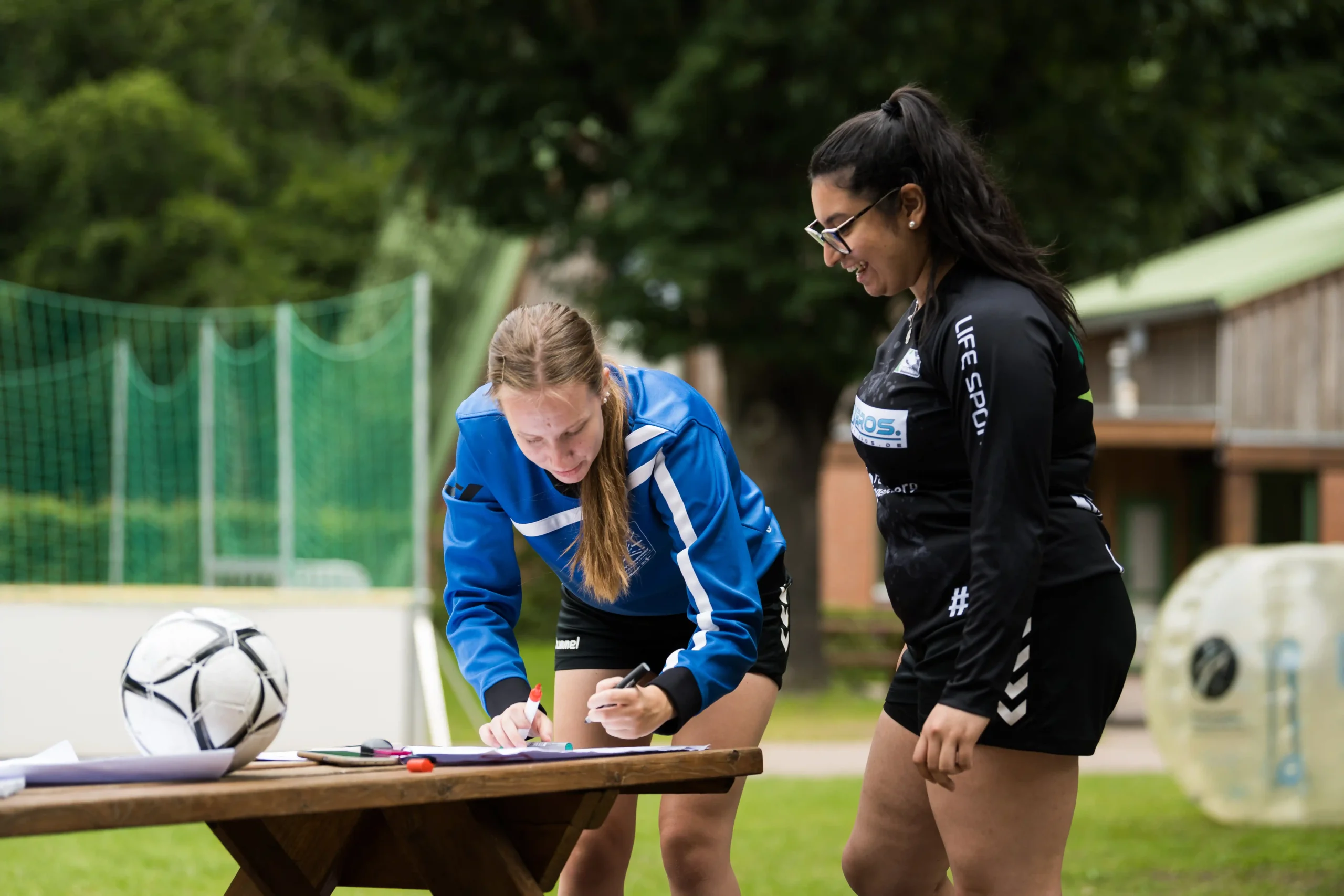 BSJ_Teambesprechung_Fussball Zwei Frauen in Sportkleidung stehen an einem Tisch im Freien; die eine schreibt etwas auf ein Blatt Papier, während die andere ihr dabei zusieht. In der Nähe sind ein Fußball und Sportausrüstung zu sehen.