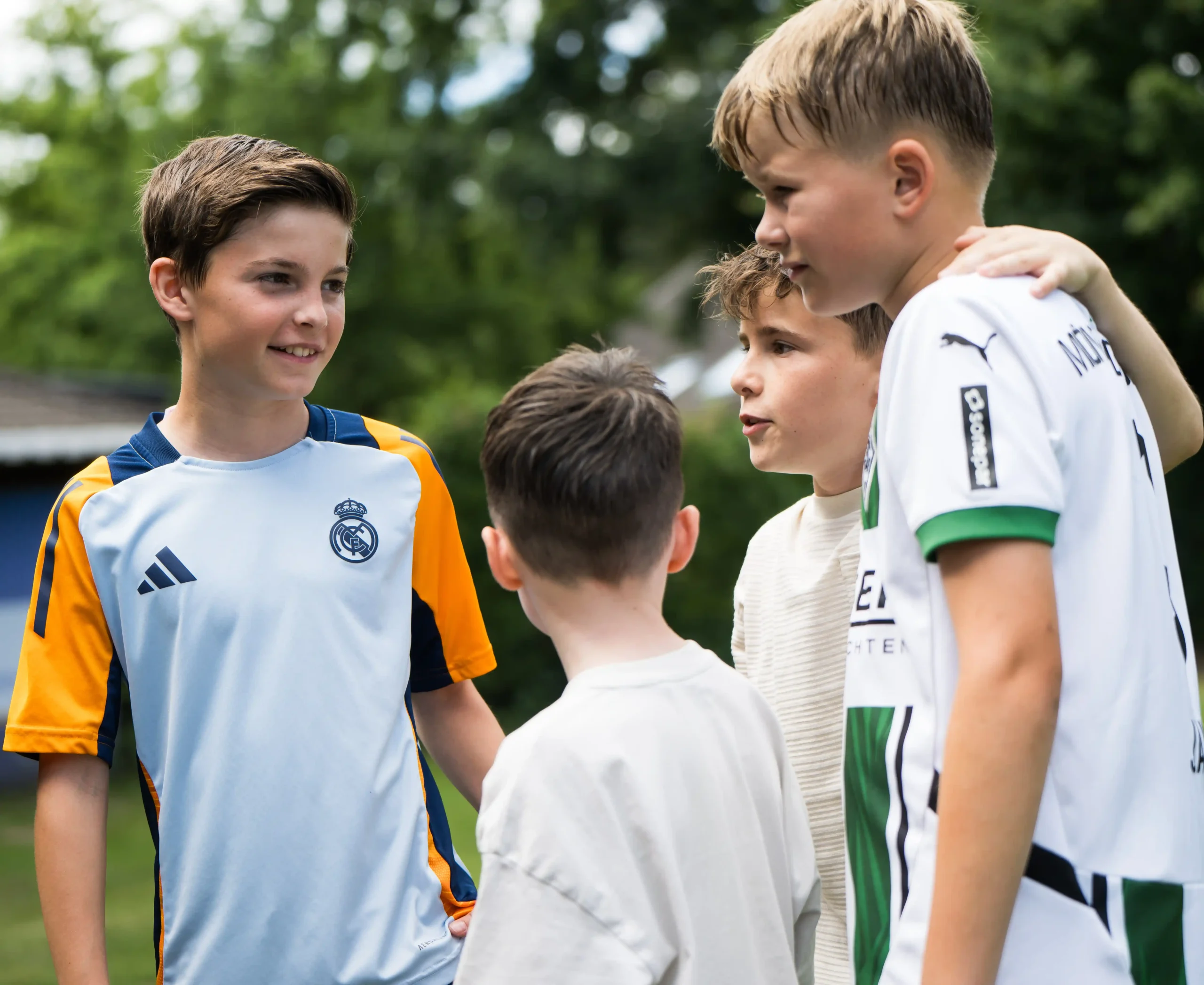 Vier Jungen stehen draußen beieinander und unterhalten sich. Zwei tragen Fußballtrikots, eines in Weiß und Grün, das andere in Hellblau mit orangefarbenen Akzenten. Im Hintergrund sind Bäume und Gras zu sehen.
