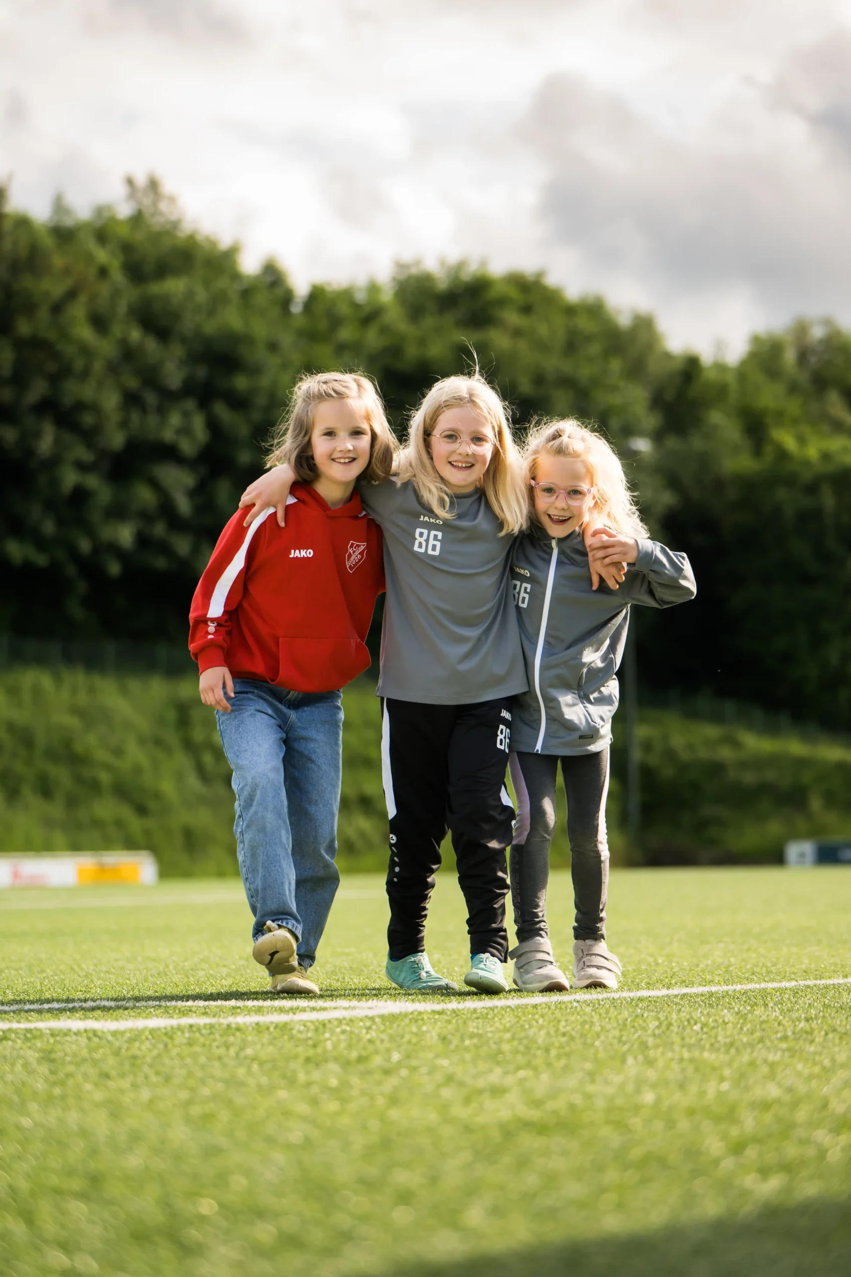 Drei lächelnde Kinder stehen Arm in Arm auf einem Fußballplatz, tragen Sportkleidung und im Hintergrund sind grüne Bäume zu sehen.