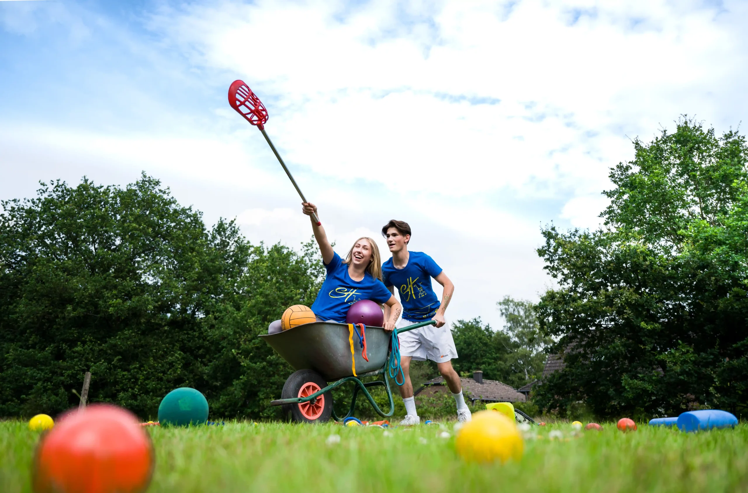 Zwei Personen in blauen Hemden posieren spielerisch mit einer Schubkarre voller Bälle auf einer Rasenfläche; eine von ihnen hält einen Lacrosse-Schläger in die Höhe, während bunte Bälle auf dem Boden verstreut liegen.