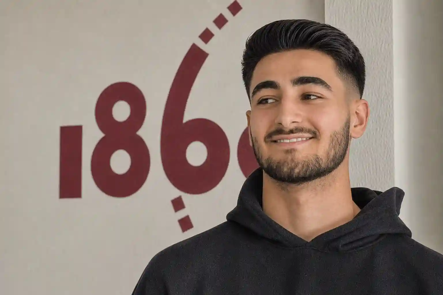 Erfahrungsberichte_FWD_Ali Ein Mann mit dunklem Haar und Bart steht lächelnd vor einer Wand, auf der in großen roten Ziffern „186“ steht.