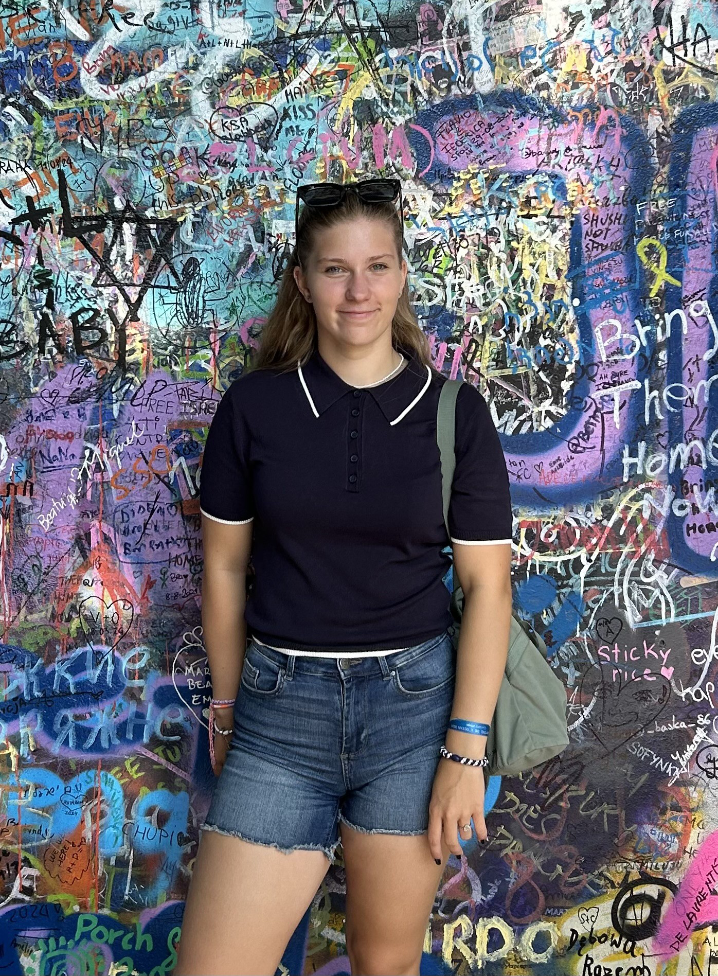 Erfahrungsberichte_FWD_Lena Kopie Eine junge Frau steht vor einer bunten, mit Graffiti übersäten Wand. Sie trägt ein dunkelblaues Shirt, Jeansshorts und eine Sonnenbrille auf dem Kopf sowie einen Rucksack über einer Schulter.