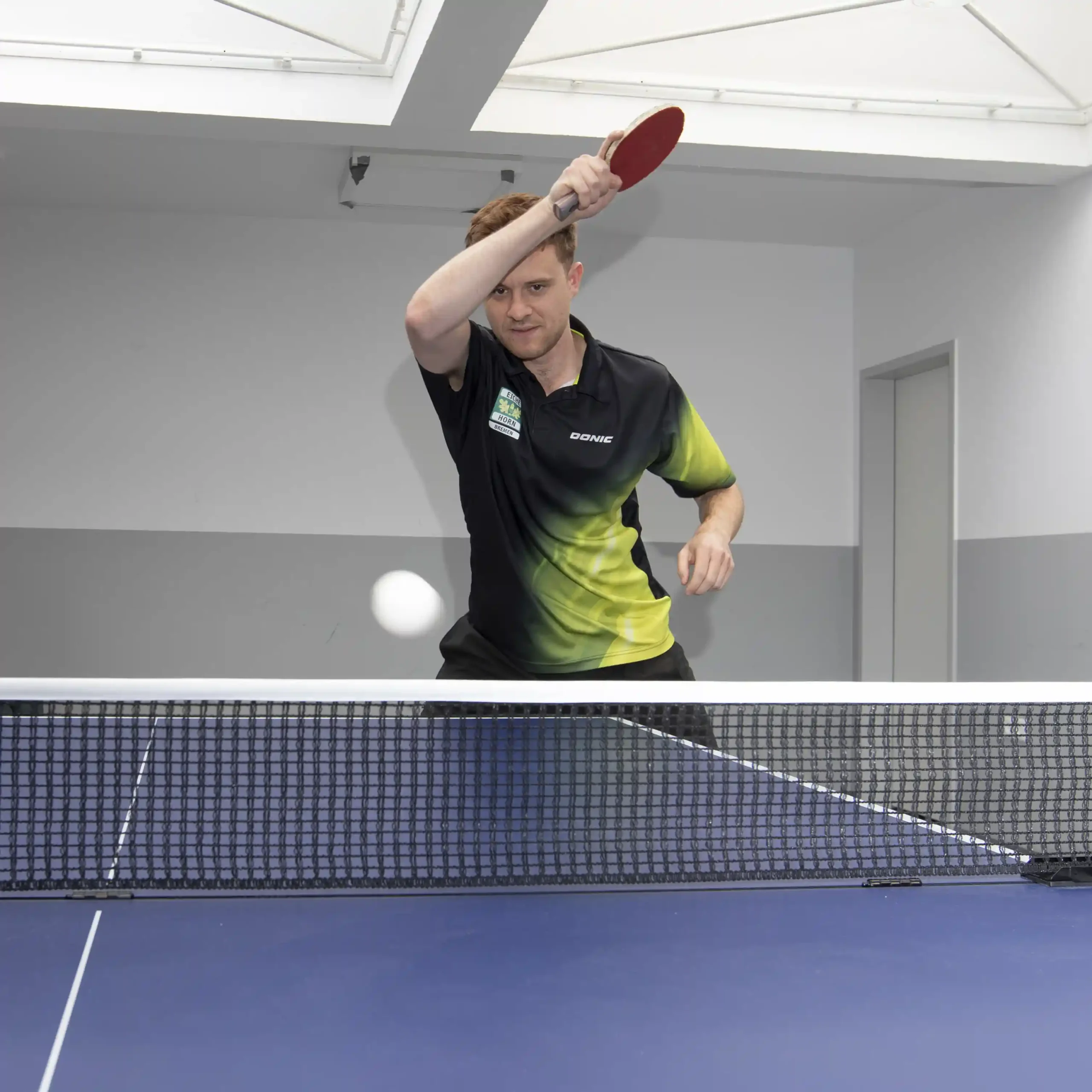 Eine Person in einem schwarz-grünen Shirt spielt in einer Halle Tischtennis und macht sich bereit, den Ball mit einem roten Schläger über einen blauen Tisch zu schlagen.