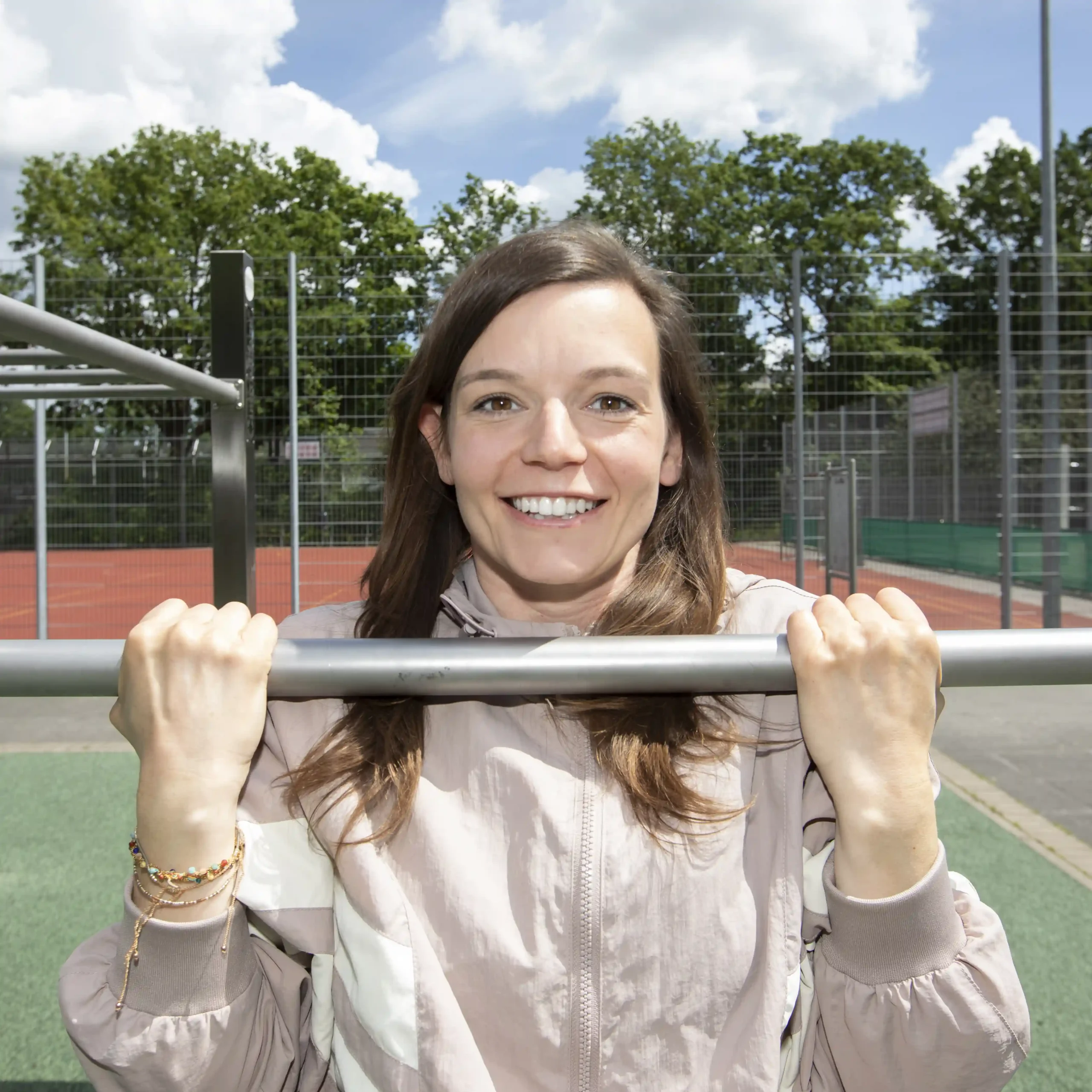 Eine Frau in einer leichten Jacke, die lächelnd an einer Outdoor-Kletterstange einen Klimmzug macht, im Hintergrund sind ein Sportplatz und Bäume zu sehen.