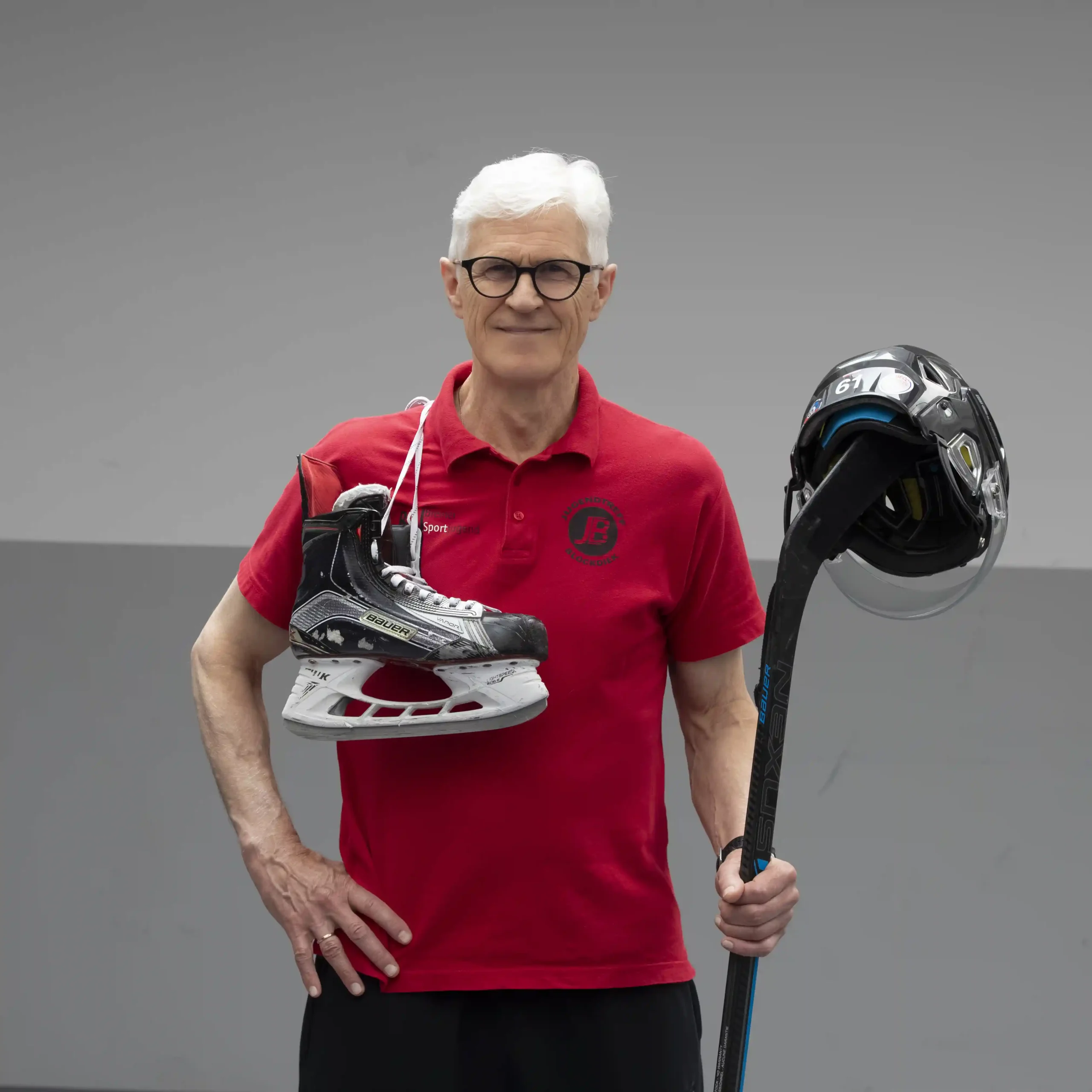 Ein älterer Mann in einem roten Poloshirt steht vor einem schlichten Hintergrund und hält einen Eishockeyschuh und einen Eishockeyschläger in der Hand, auf dem ein Helm liegt.