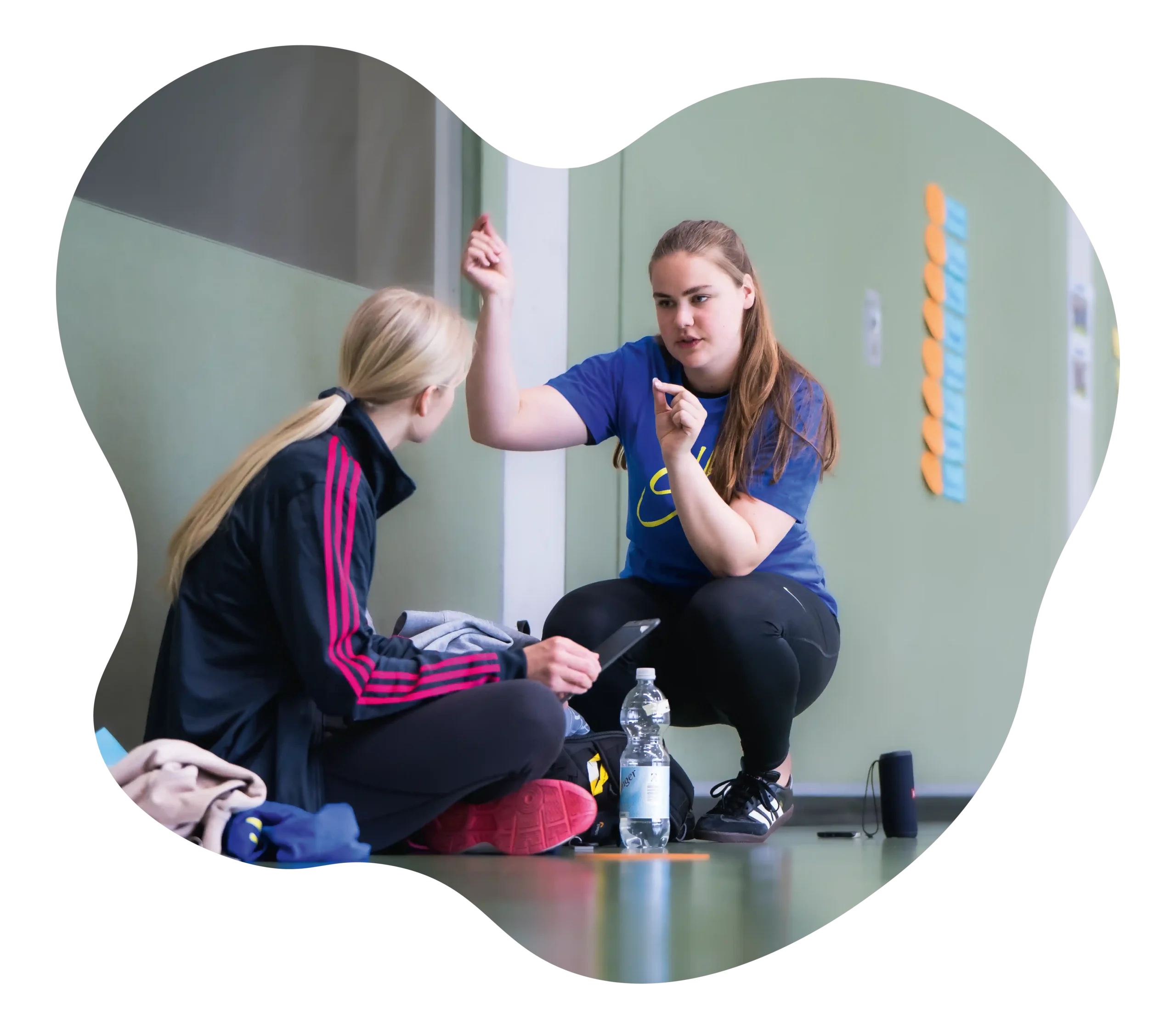 Zwei Frauen sitzen auf dem Boden einer Sporthalle; die eine gestikuliert beim Sprechen mit dem Arm, während die andere zuhört. In der Nähe stehen eine Wasserflasche und Sporttaschen, im Hintergrund sind Haftnotizen an der Wand zu sehen.