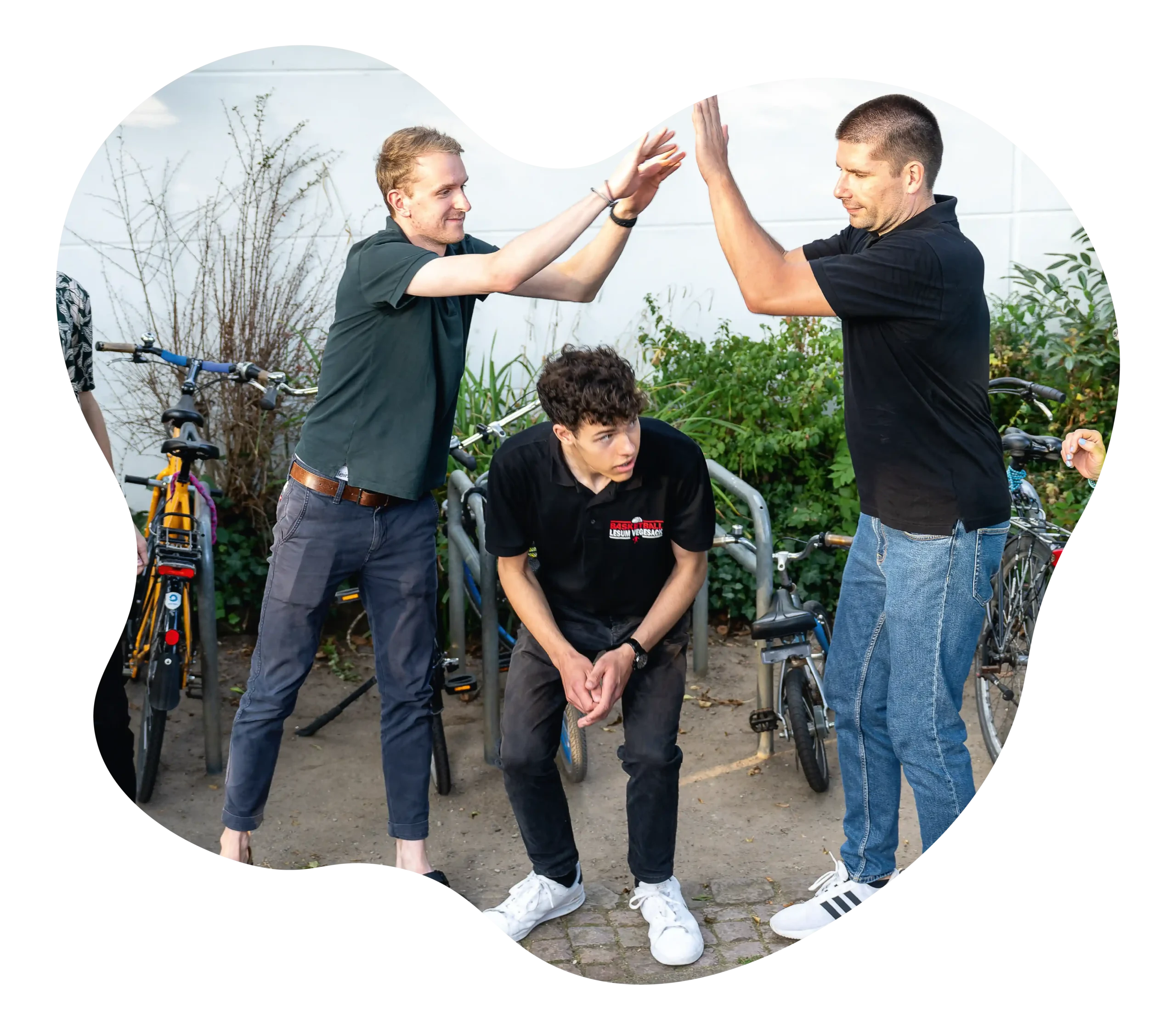 Zwei stehende Männer geben sich ein High Five, während ein dritter Mann zwischen ihnen sitzt; im Hintergrund sind Fahrräder und Grünflächen zu sehen.