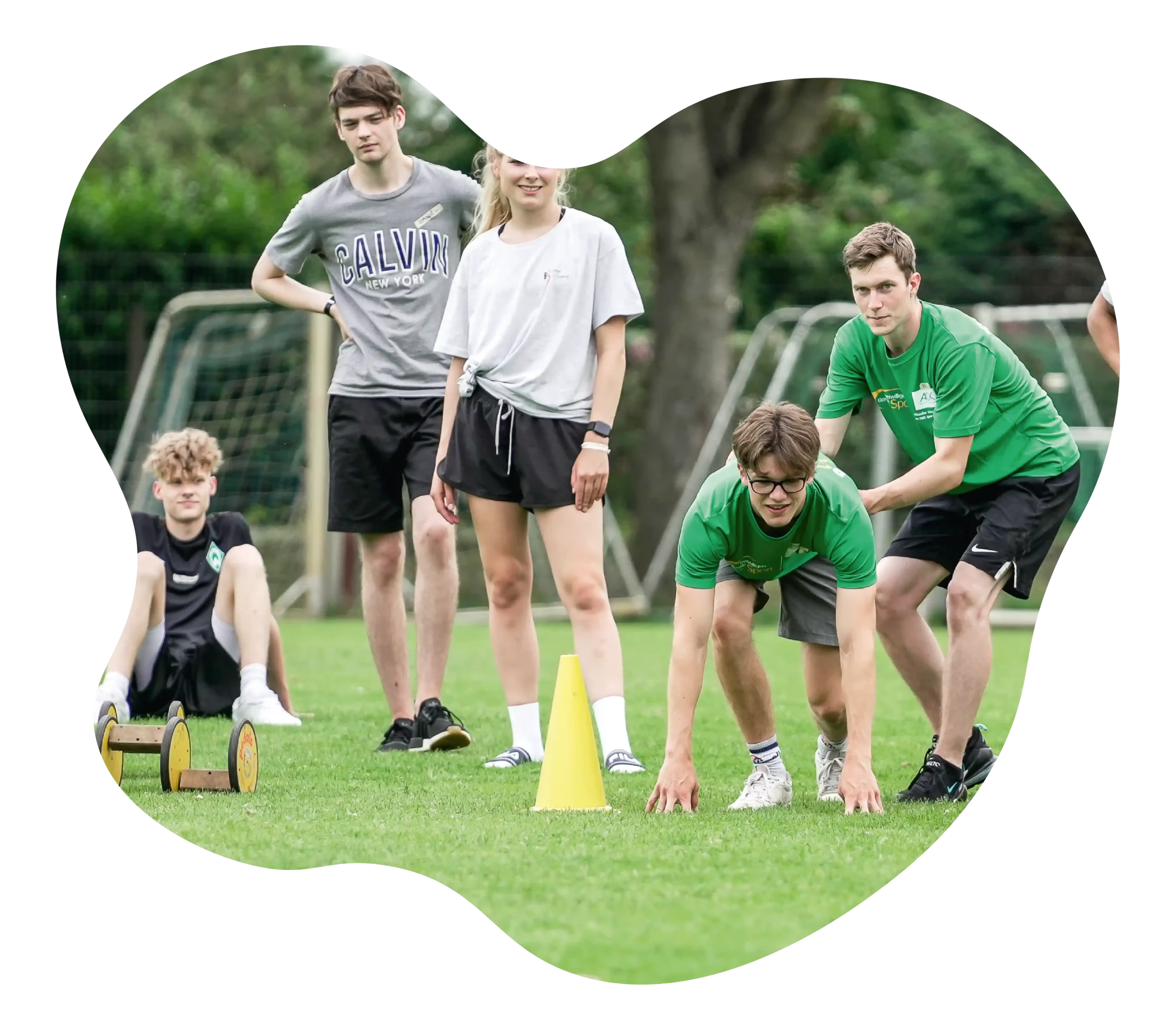 Fünf Jugendliche stehen und hocken auf einer Wiese und nehmen an einer Aktivität im Freien teil, bei der Pylonen und ein kleiner Holzkarren zum Einsatz kommen. Im Hintergrund sind Fußballtore zu sehen.