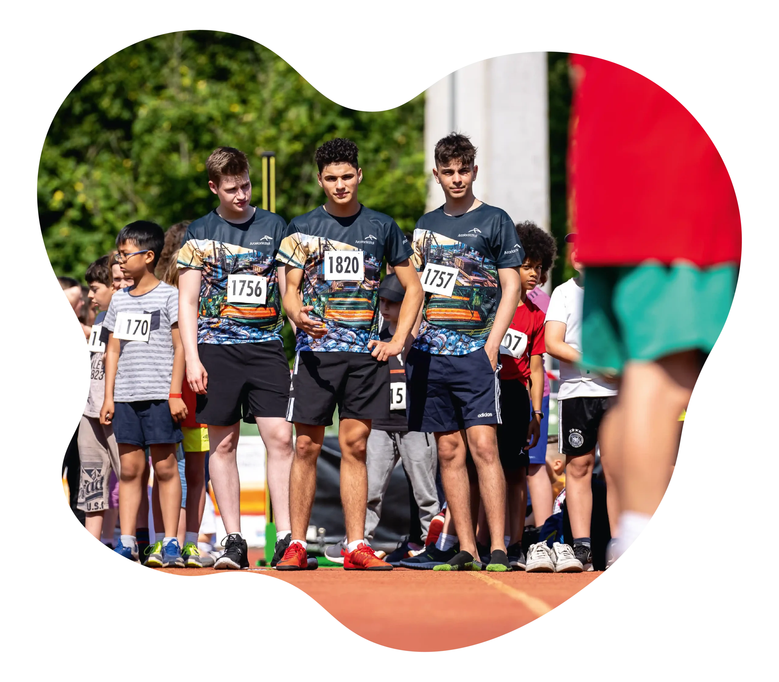 Mehrere Läufer mit Startnummern stehen an der Startlinie einer Leichtathletikveranstaltung im Freien und bereiten sich auf ein Rennen vor. Im Vordergrund steht eine Person in rot und grün.