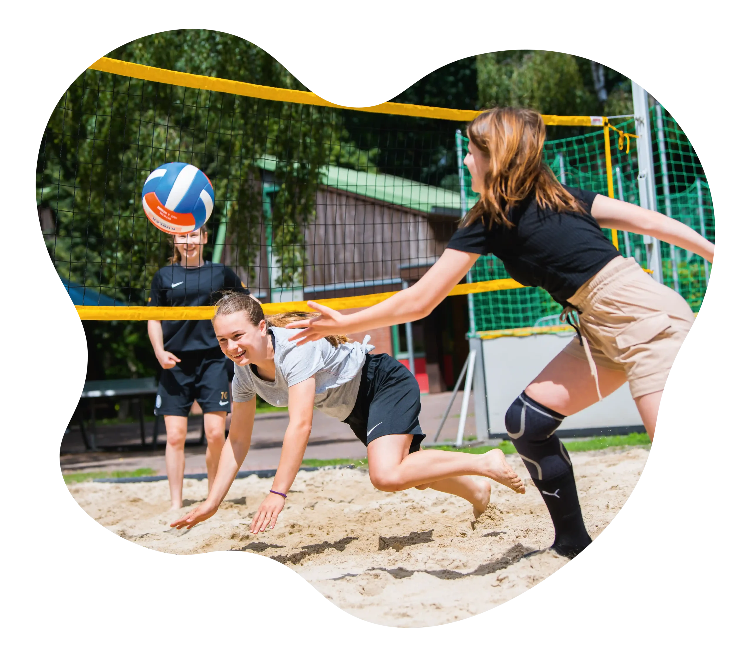 Drei Personen spielen Beachvolleyball; ein Spieler im grauen T-Shirt streckt sich nach dem Ball, während ein anderer in einem schwarzen T-Shirt und hellbraunen Shorts nach vorne läuft. Ein dritter Spieler steht im Hintergrund.